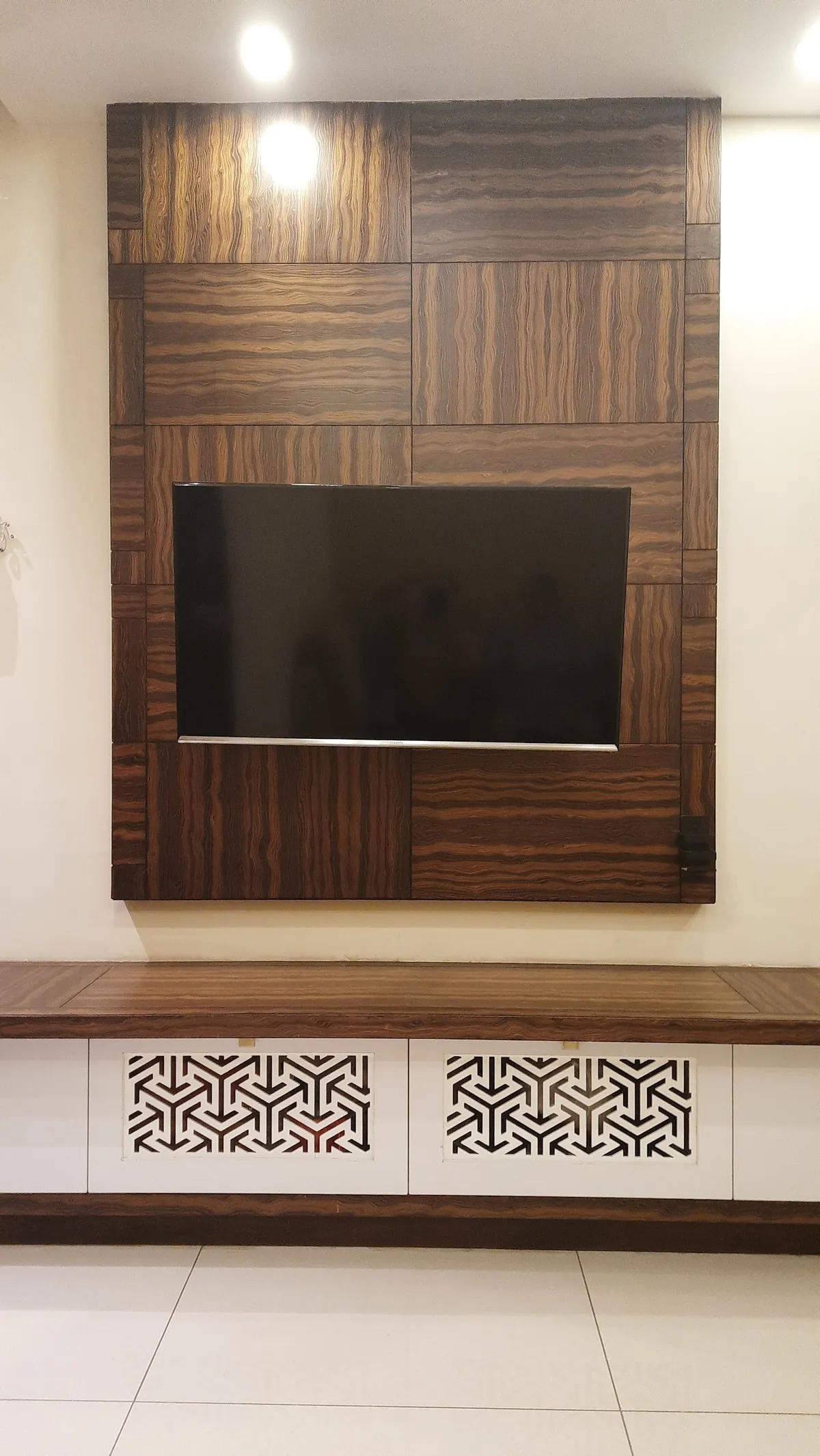 Living room TV feature wall made of patchwork wood-veneer panels with a mounted TV, above a floating console with geometric jaali-pattern cutwork inserts.