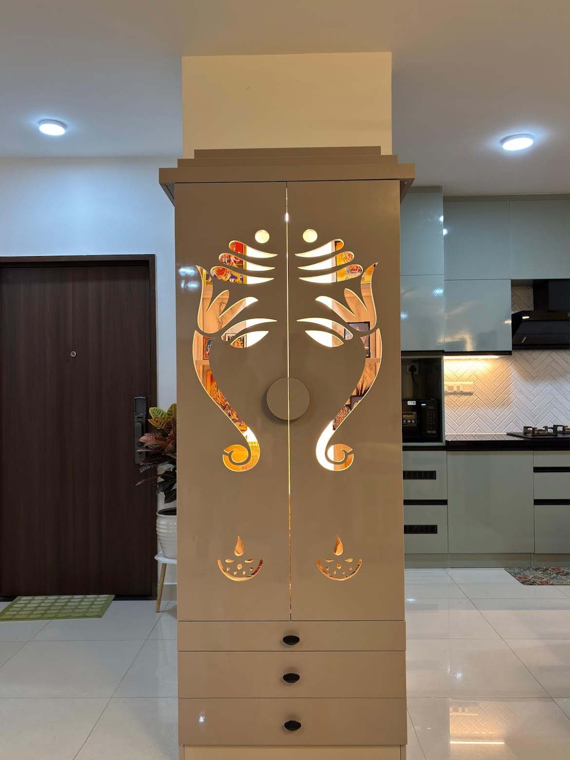 Standalone pooja cabinet with backlit laser-cut Ganesh-motif doors and diya cutouts, positioned near the entrance with a glimpse of a mint-green kitchen behind.