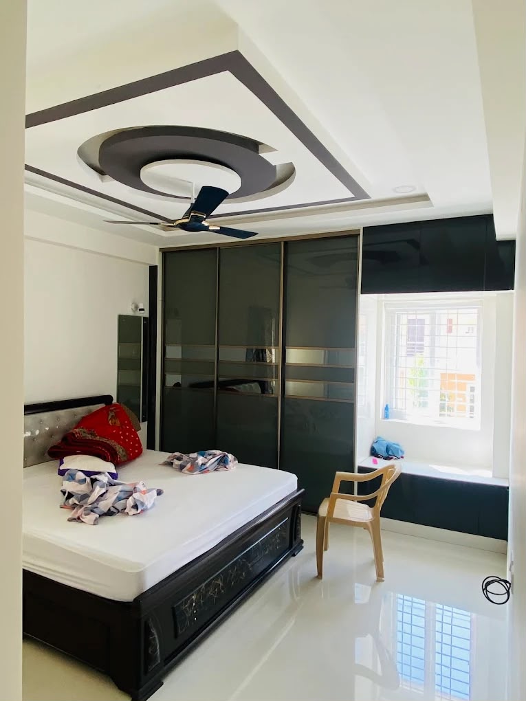 A bedroom with a circular POP false-ceiling ring around the fan, a black glossy sliding wardrobe with mirror-strip inlays, and a carved dark-wood bed frame