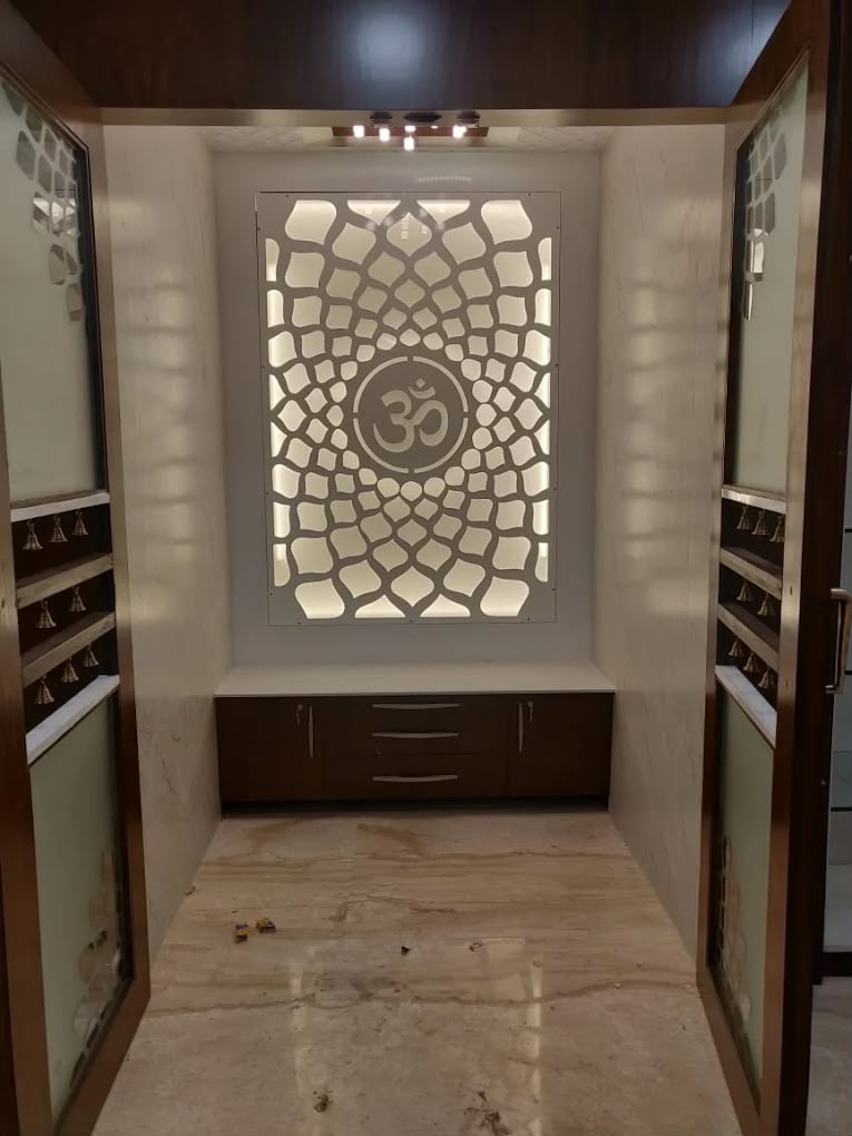 Pooja alcove with a backlit CNC-cut jaali panel featuring an Om motif above a wood-and-marble cabinet, flanked by mirrored wardrobe doors with hanging bells.