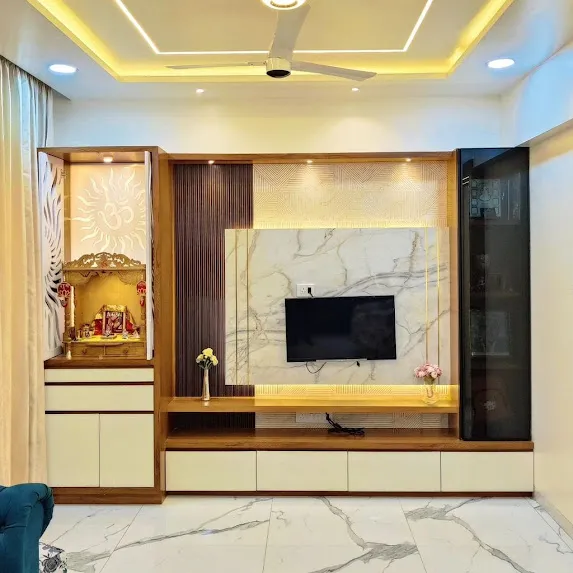 Living-room feature wall pairing a backlit pooja niche with a carved sun motif and marble idol platform with a marble-slab TV panel between fluted teak columns.