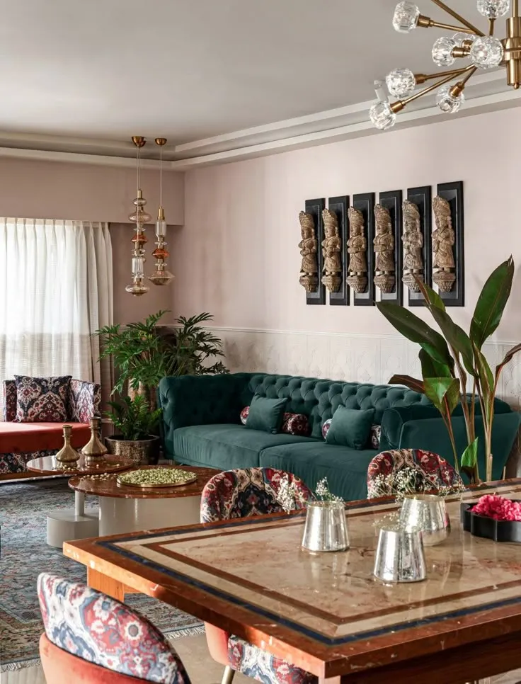 Blush-walled living room with an emerald velvet chesterfield under six carved wooden deity panels in black frames, a brass sputnik chandelier overhead