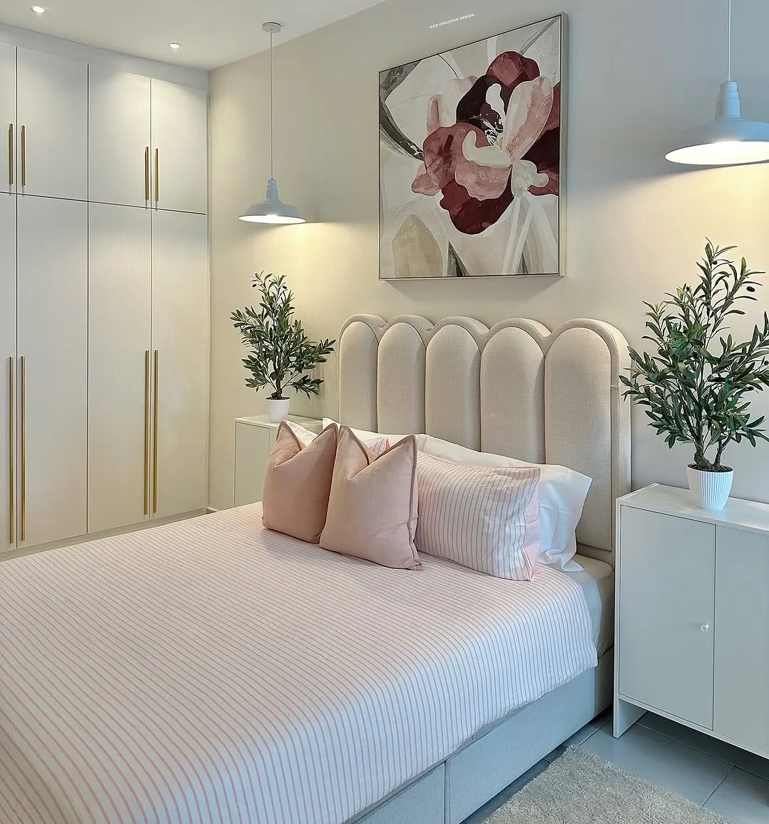 Bedroom with a cream scalloped-arch upholstered headboard and blush striped bedding, flanked by two white dome pendants and potted olive trees
