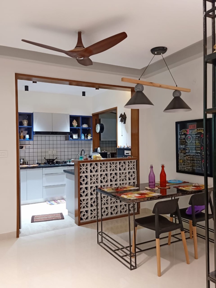 Open dining nook with a black-metal-framed glass dining table under twin cone pendant lights, a jaali partition screening the kitchen behind