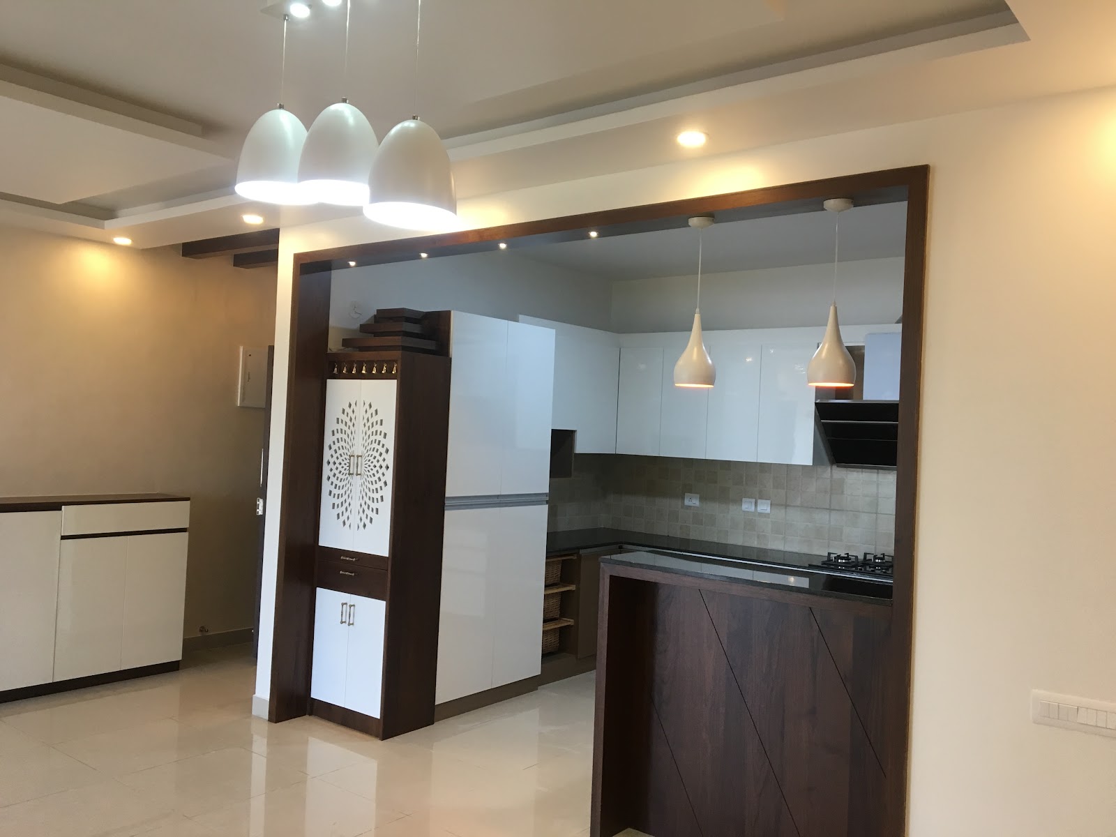 Open kitchen framed by a dark walnut archway with a built-in jaali pooja unit and white gloss cabinetry beyond
