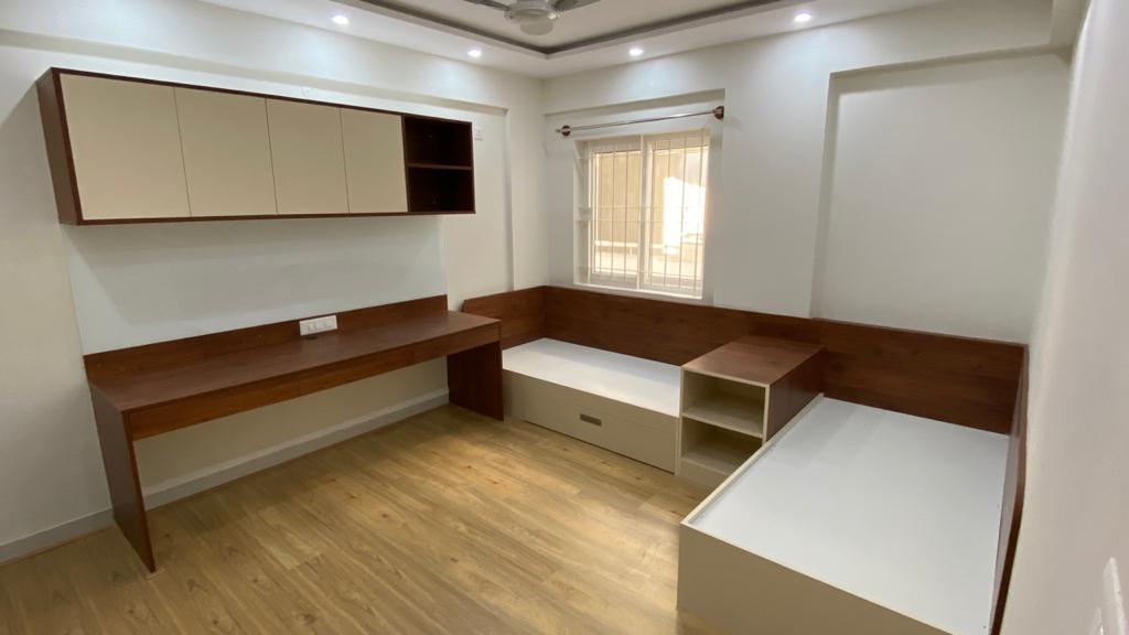 Children's bedroom with twin built-in platform beds along two walls, a matching study desk, and an overhead storage cabinet, in maroon and cream laminate.