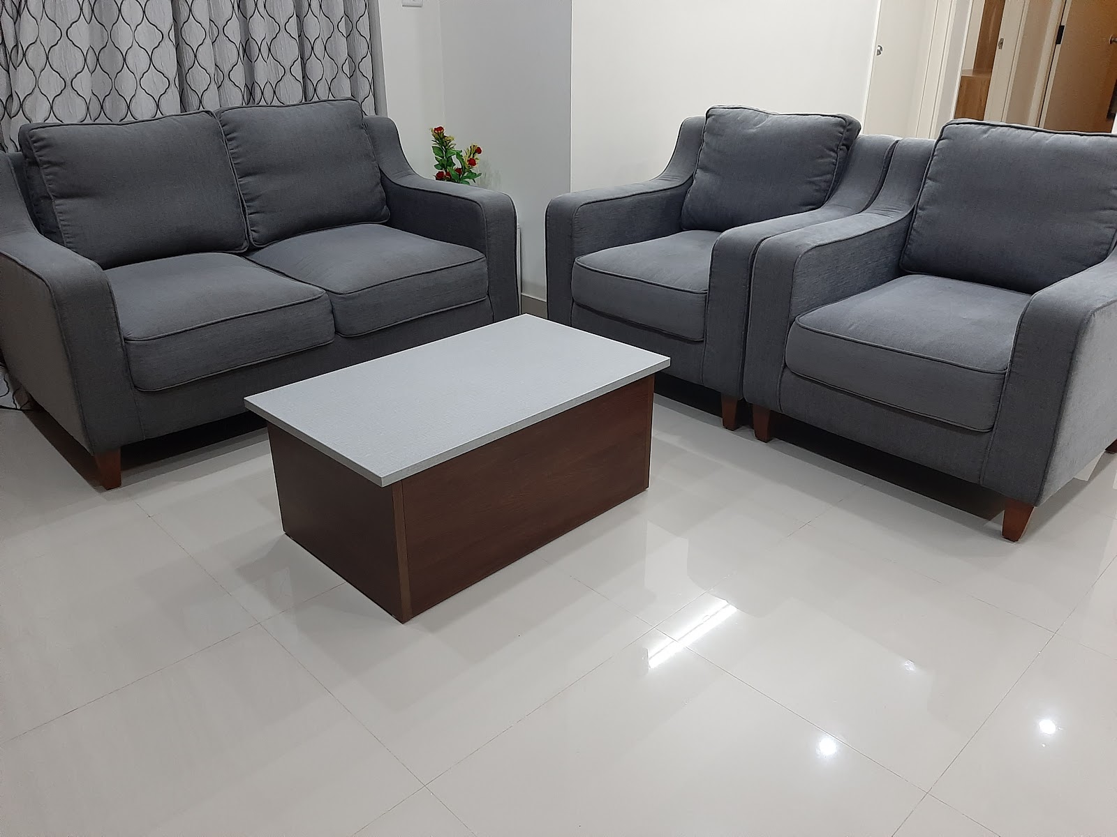 Living room with a grey fabric three-piece sofa set arranged around a wood-and-white coffee table, patterned sheer curtains, and plain white walls and flooring.