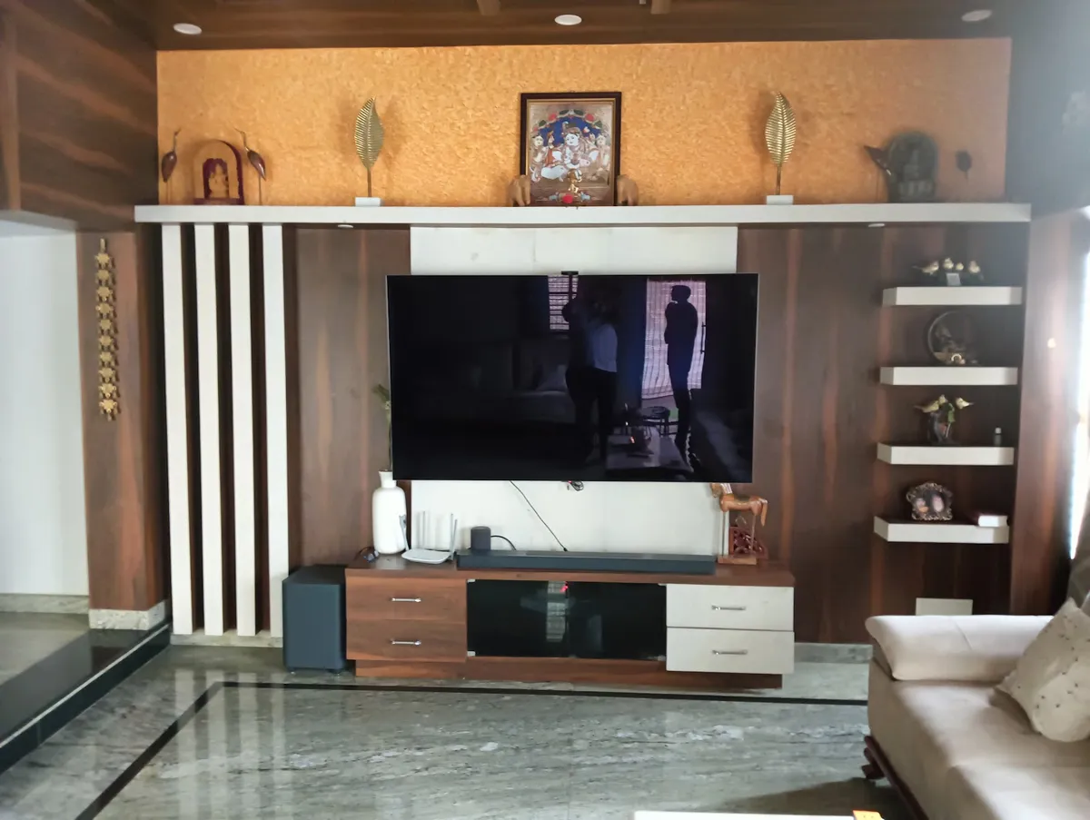 Living-room TV wall in dark walnut veneer with white fluted battens to the left, an ochre textured band above holding a framed Krishna painting and brass leaf