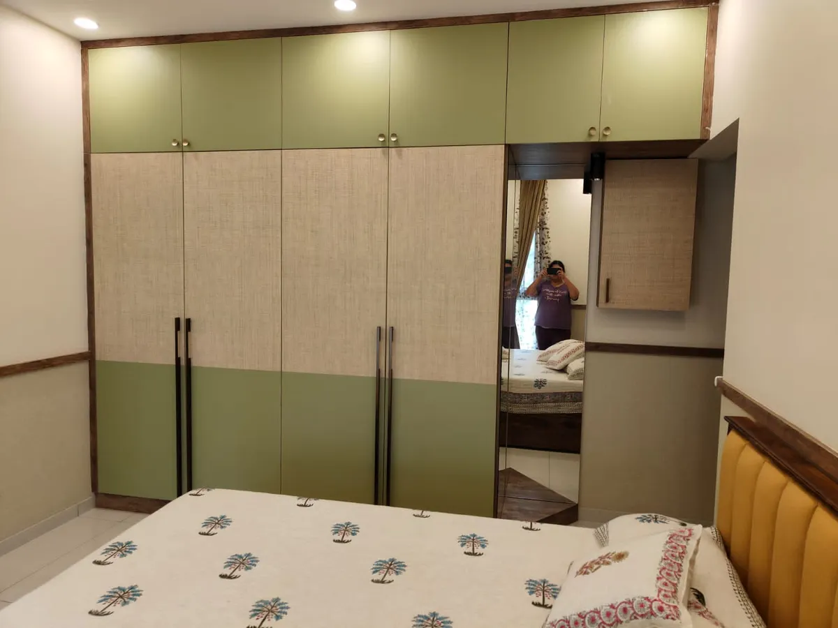 Bedroom wardrobe wall in sage-green and jute-textured laminate with separate lofts above and a full-height mirror panel at the end