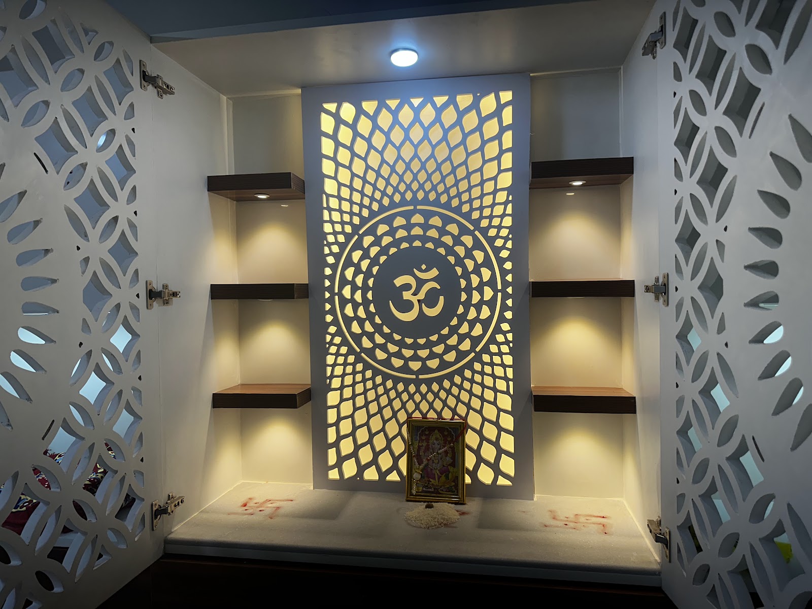 Interior of a pooja unit with CNC-cut floral jaali doors open to reveal a backlit Om-motif panel, marble shelf with swastik drawn in kumkum