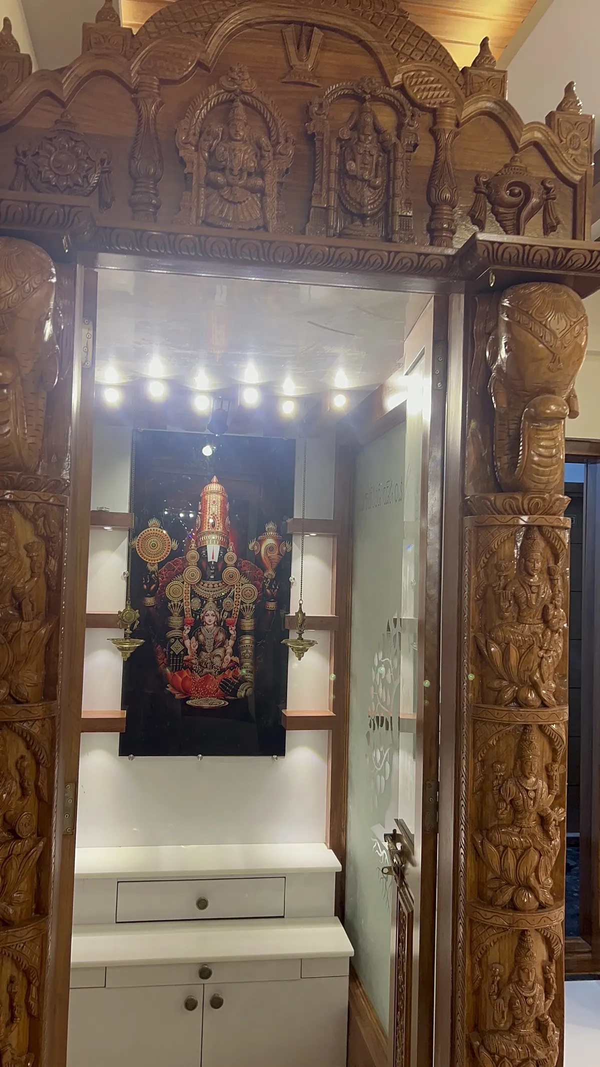 Elaborate hand-carved wood pooja mandap with elephant-pillar carvings and deity relief work, a backlit framed deity portrait
