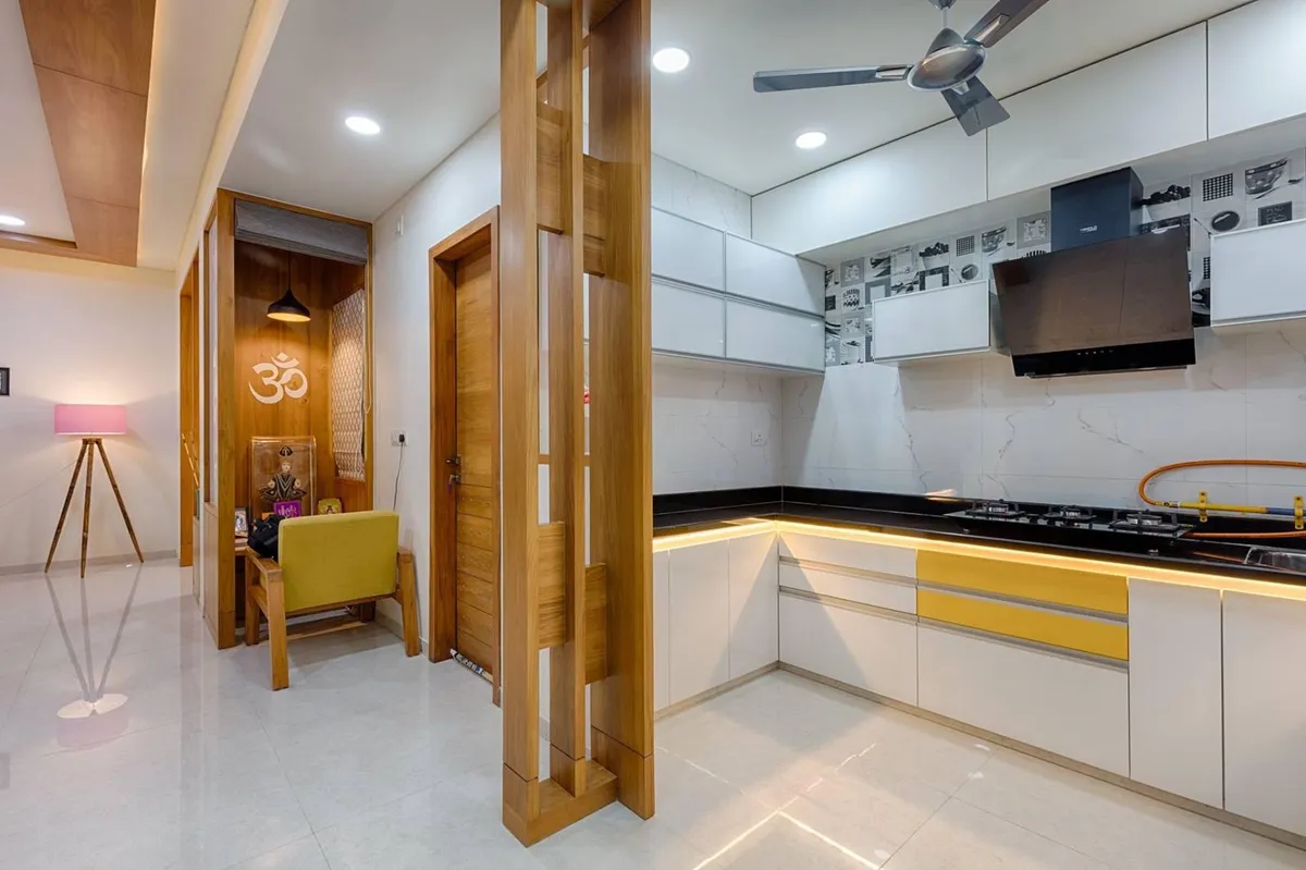 Open kitchen with mustard-yellow accent shutters and a black granite counter, opening onto a wood-panelled pooja nook with an Om wall motif.