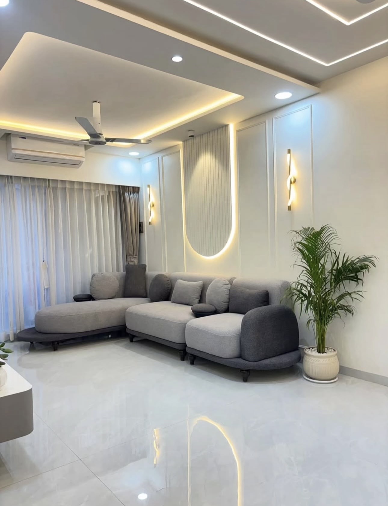 Living room with a curved grey modular sofa facing a backlit fluted wall panel flanked by wall sconces, under a cove-lit stepped ceiling.