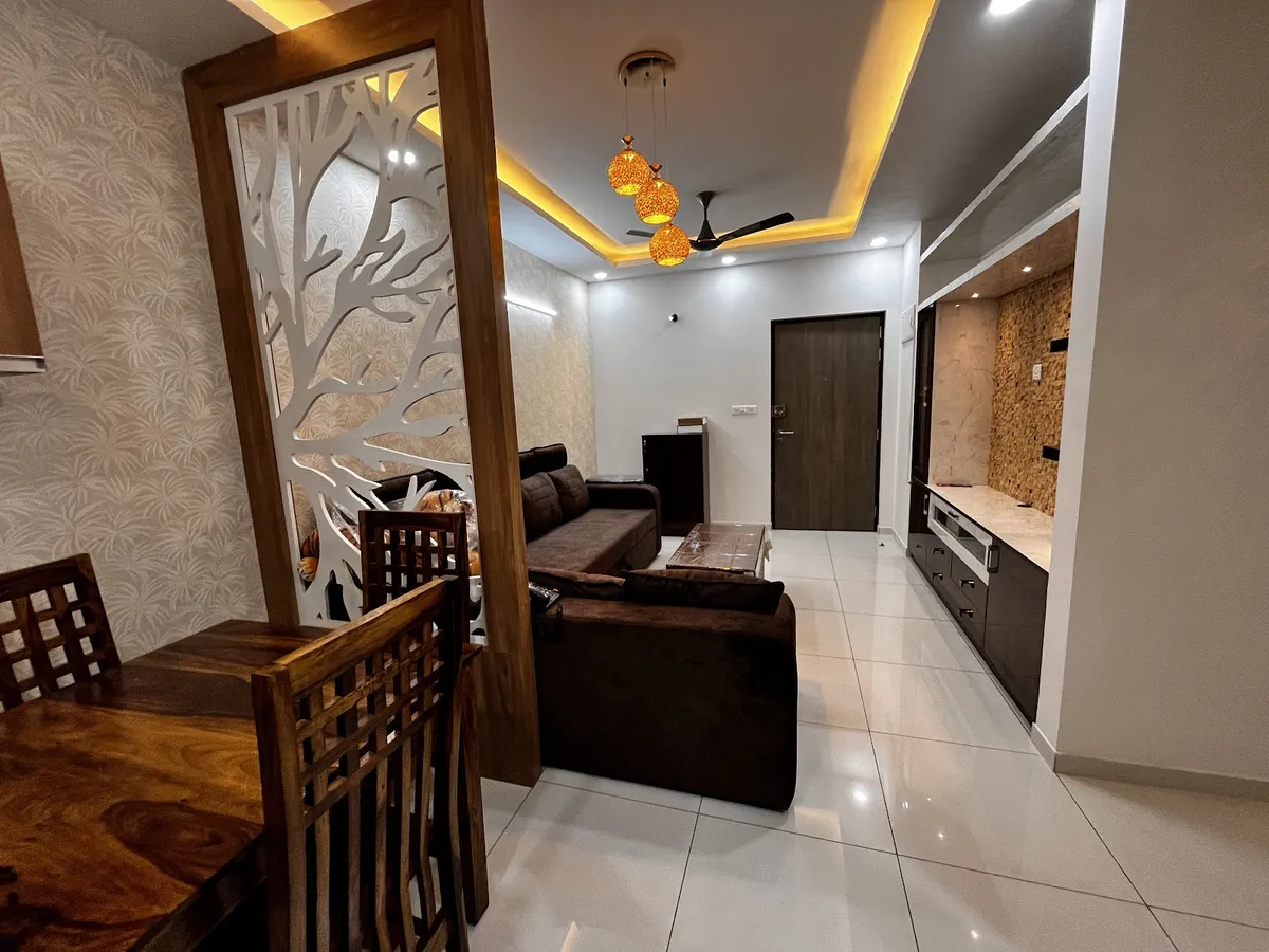 Open-plan dining and living area divided by a tree-branch cutout wood partition, with amber cluster pendant lights, a brown sofa, and a stone-clad TV wall.