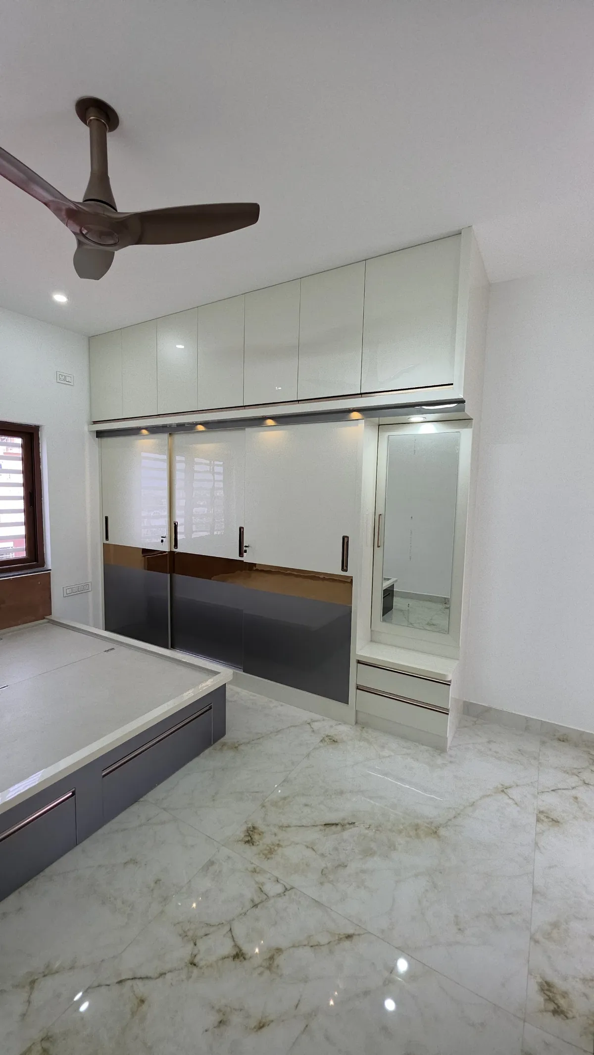 Bedroom with a floor-to-ceiling sliding wardrobe in grey and bronze-accented glossy shutters, an attached mirrored dresser with drawers, and marble flooring