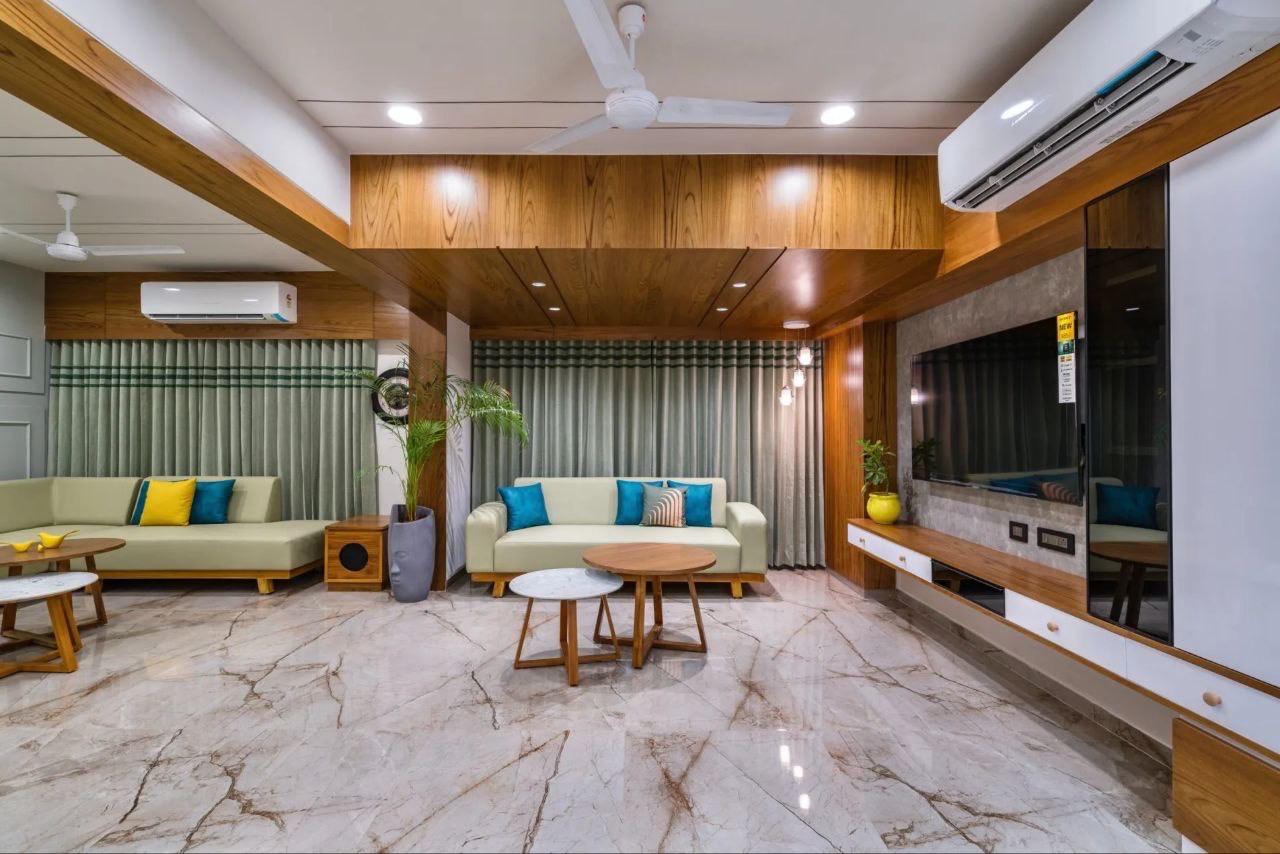 An open living room with a wood-paneled ceiling bulkhead, a stone-clad TV wall, and olive-green sofas with teal accent cushions on a veined marble floor.