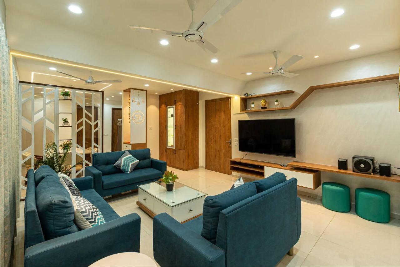 Living room with three teal sofas around a white glass coffee table, an angled floating wood TV shelf, and a white geometric jaali room divider.