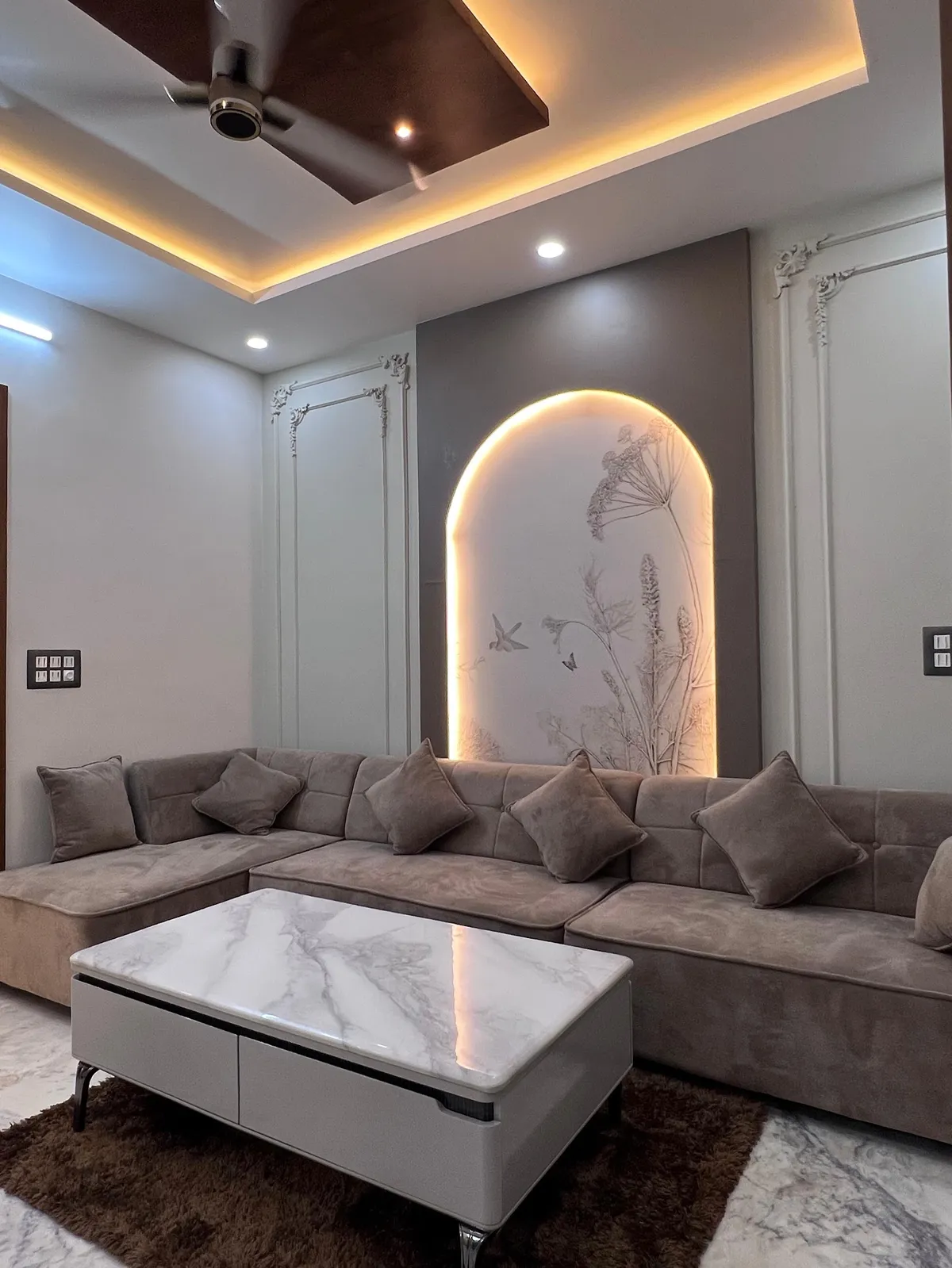 A living room with a backlit arched wall panel featuring botanical line-art, flanked by ornate wall moulding, a grey tufted L-shaped sofa