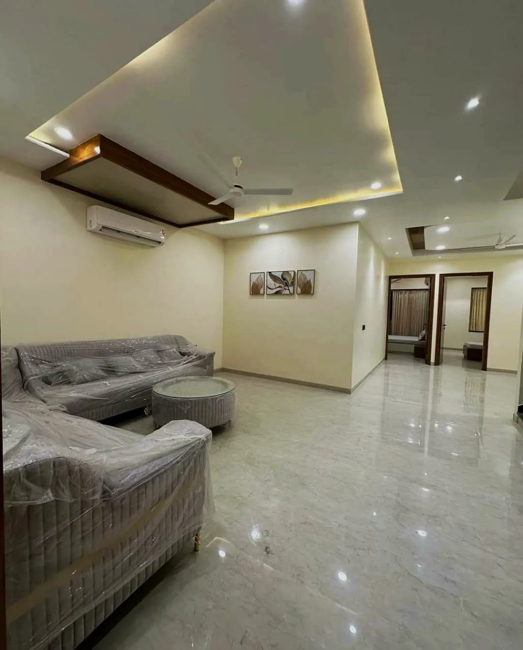 Living room of a just-handed-over flat with the grey L-sofa still in its factory plastic wrap, a fluted round coffee table
