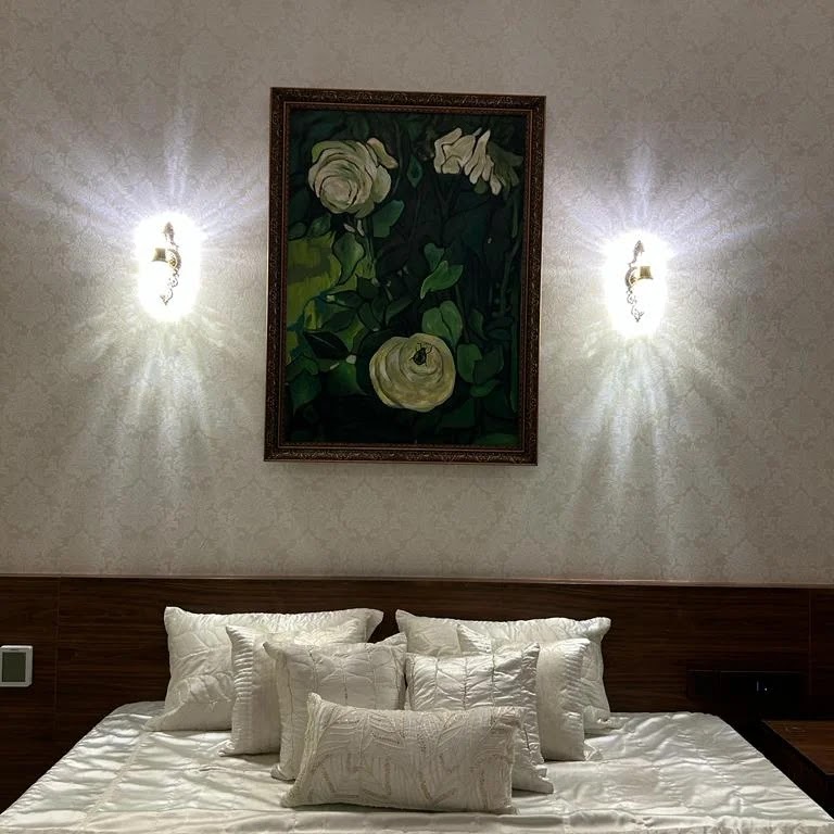 A master bedroom with damask wallpaper, a wood-panelled headboard, symmetrical wall sconces flanking a framed floral painting, and layered white pillows