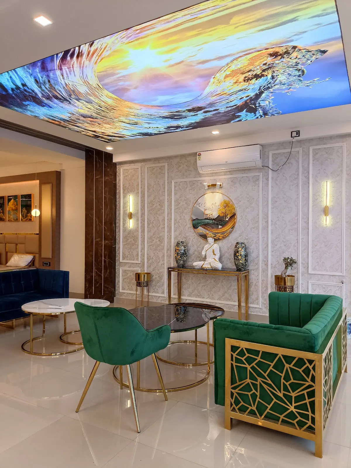 Backlit stretch-ceiling print of a breaking wave over an emerald velvet sofa with a gold jaali-pattern frame, a Buddha idol and twin jars on a brass console