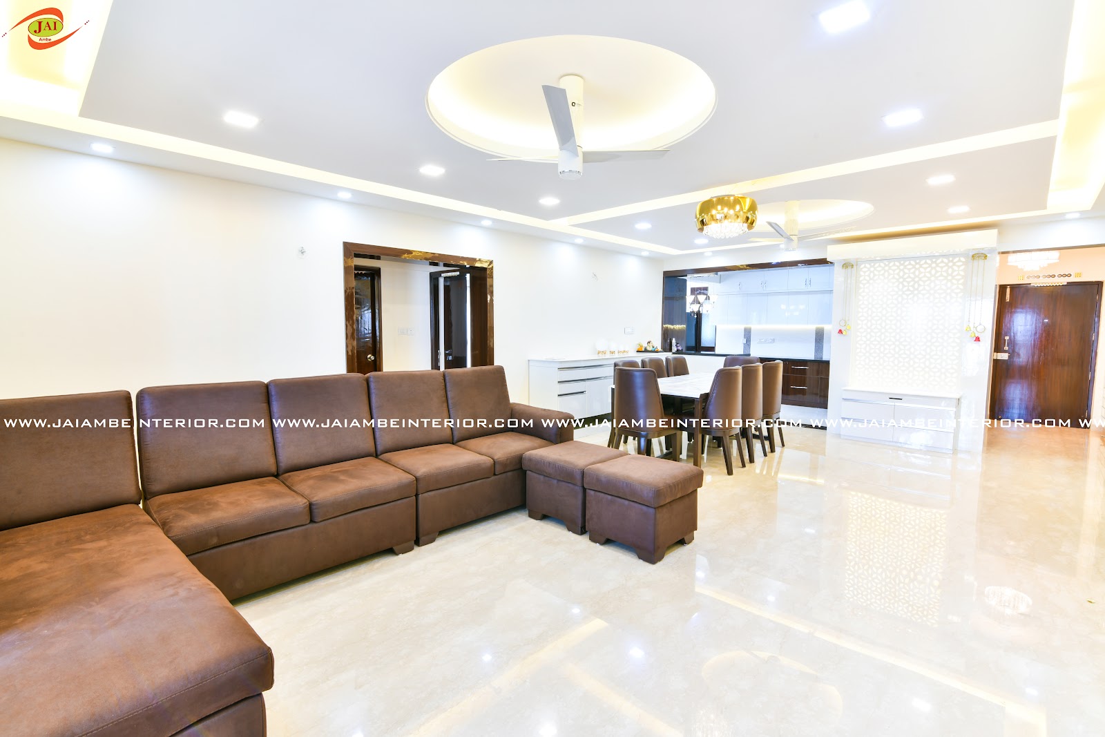 Open-plan apartment shot showing a brown sectional sofa in the foreground, a dining table under a crystal pendant, a kitchen counter