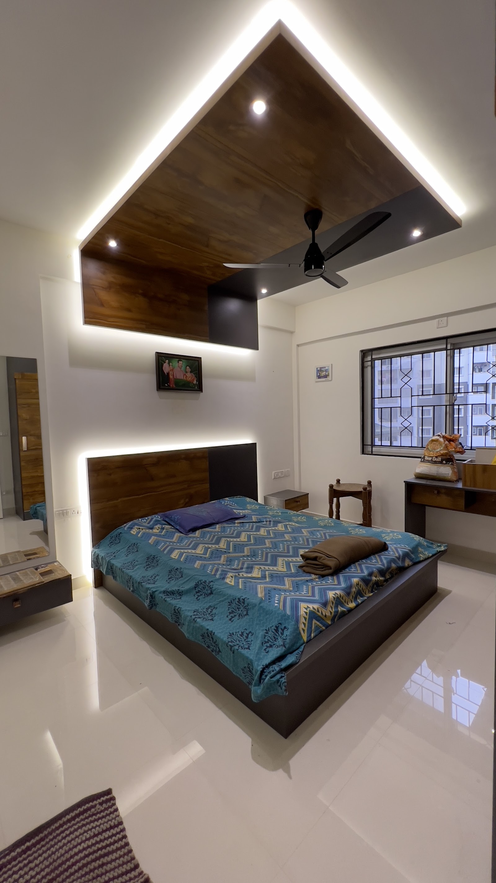 Bedroom with a dramatic wood-clad false-ceiling drop with LED cove lighting above a wood-and-black headboard bed, a small wall-mounted study desk