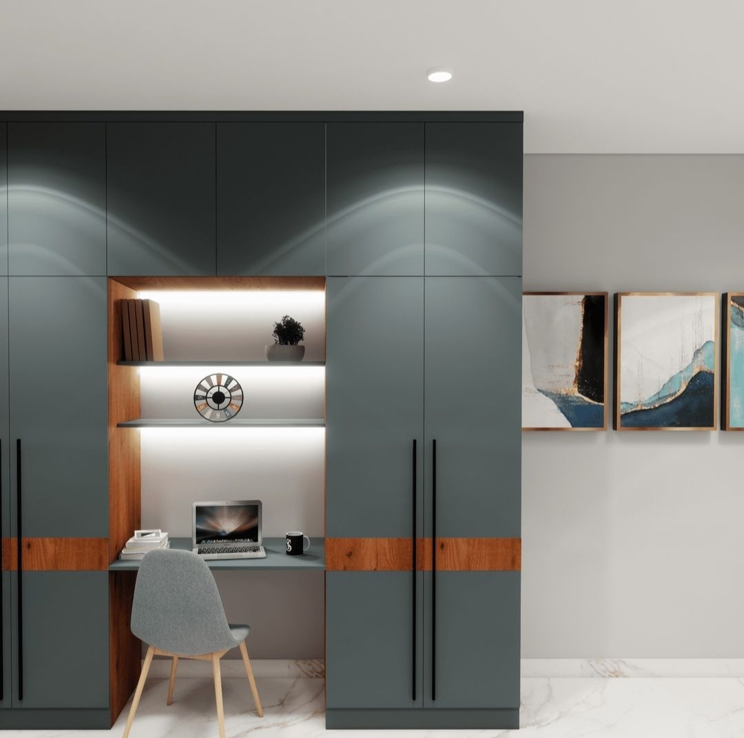 Render of a built-in study nook with lit open shelving inside a dark teal-grey wardrobe wall, a laptop desk, and a grey accent chair.