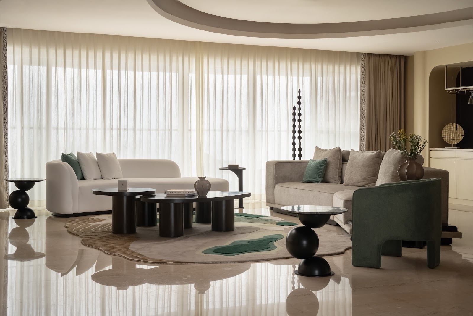 A living room with sculptural black pedestal coffee and side tables on an organic-shaped rug, floor-to-ceiling sheer curtains, and cream and sage-green sofas.