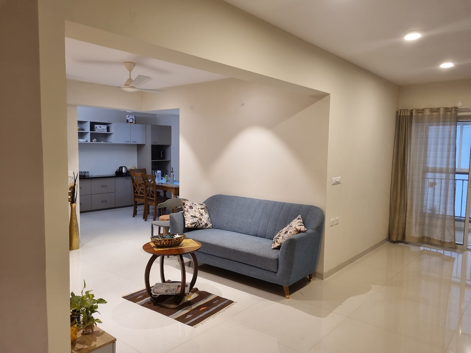 Living room with a blue channel-tufted sofa and a round wood side table, opening onto a kitchen and dining area through an archway.