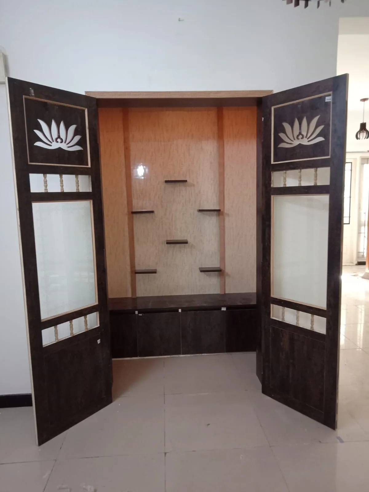 Newly fitted pooja mandir with lotus-cutout and frosted-glass folding shutters swung open to staggered display shelves over a closed storage base