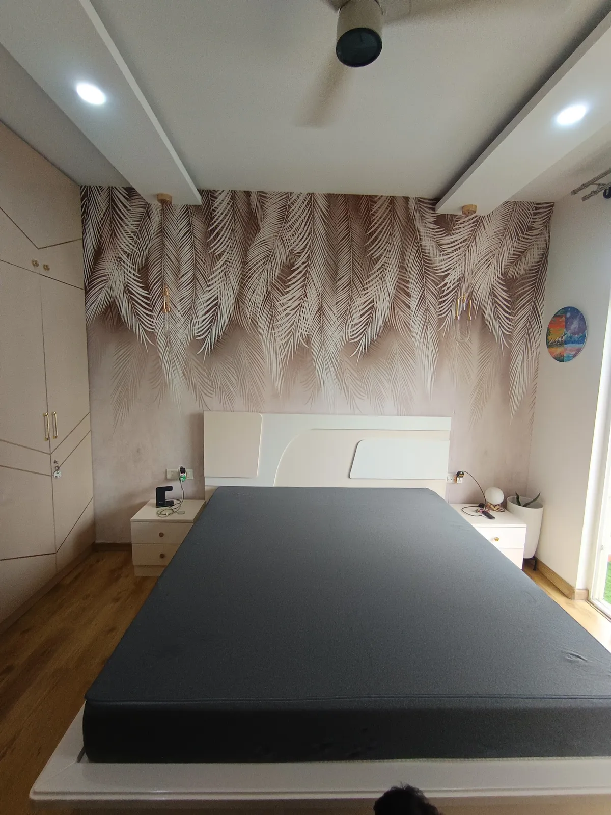 Photograph of a bedroom with a blush palm-frond mural covering the headboard wall, slim brass pendant drops hanging either side of a white curved headboard