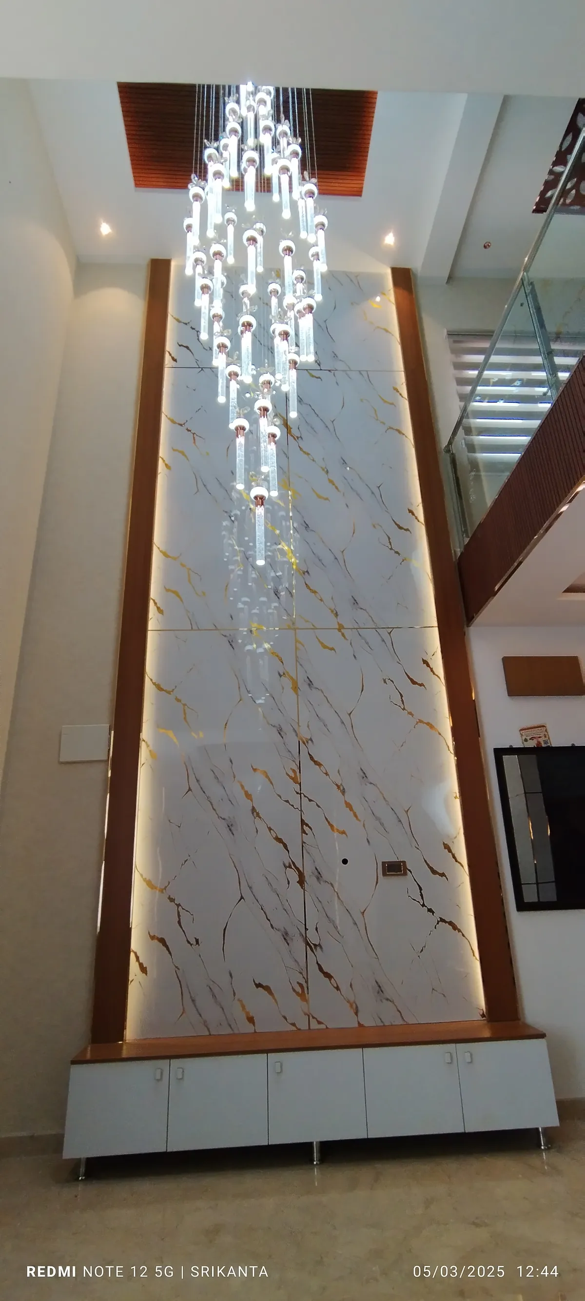 Double-height entrance foyer with a backlit gold-veined marble feature wall, a long cascading crystal chandelier, and a white console below