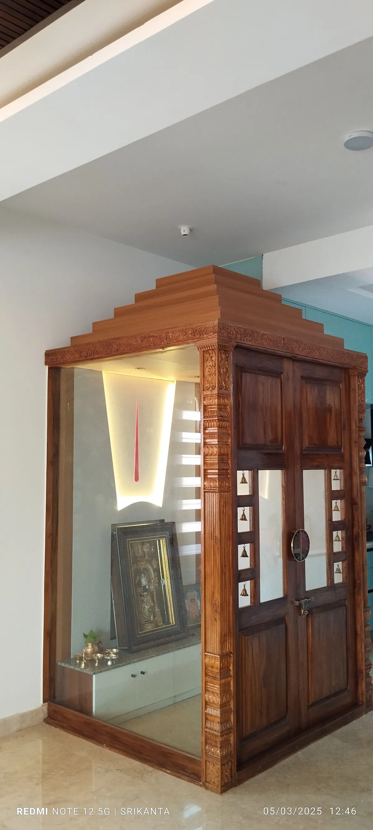 Freestanding glass-walled mandir cabin in a corner of the home, with a stepped teak gopuram cornice
