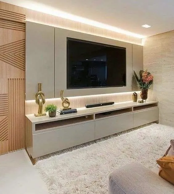 A taupe TV console wall with a warm backlit panel, a geometric carved-wood sliding door, and a plush cream shag rug in front.