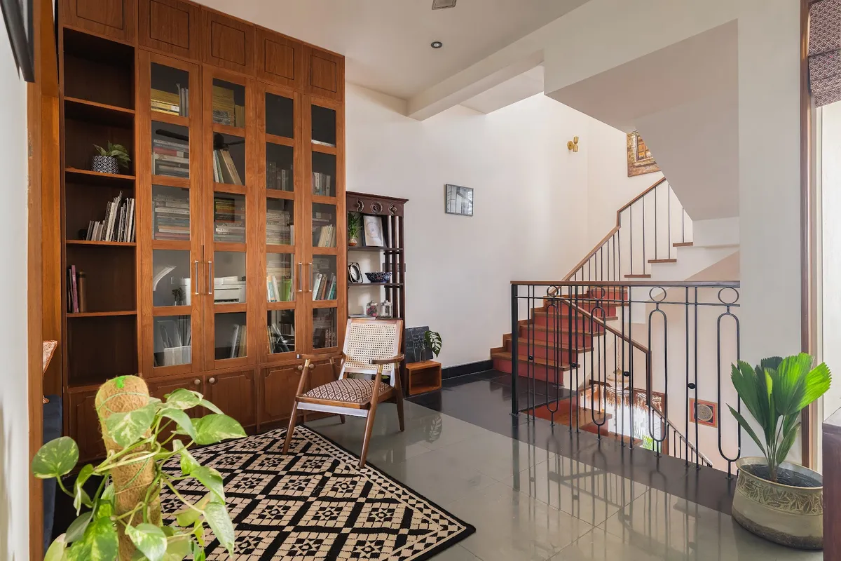 Reading nook at the foot of a staircase, with a full-height glass-front wood bookcase, a cane-back accent chair, and a black-and-white patterned dhurrie rug.