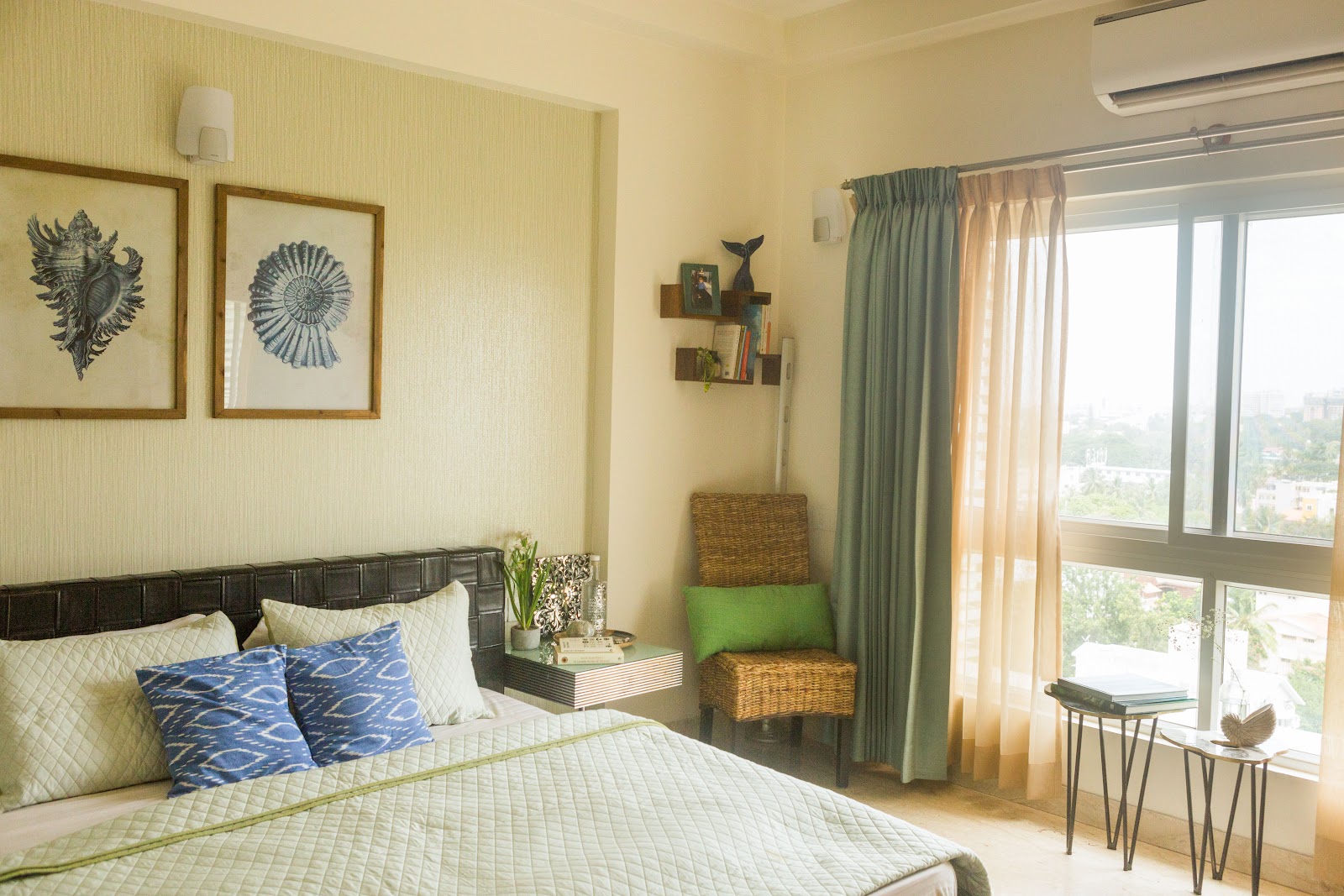 Sage-green bedroom styled with framed seashell prints, an indigo ikat-cushioned bed, a woven rattan accent chair by the window, and floating nightstands.