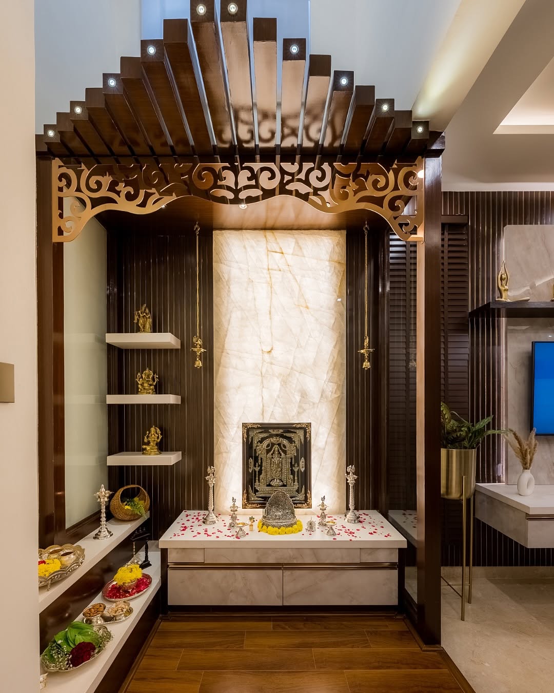 A pooja room with a fan-shaped carved wood arch trimmed in brass filigree, a backlit onyx stone niche