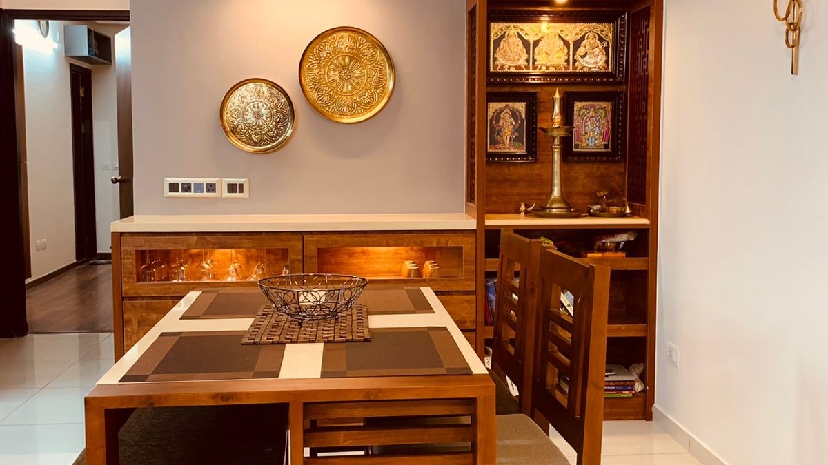 Dining area with a wood table, brass decorative wall plates, and an adjoining built-in pooja unit with Tanjore-style deity paintings.