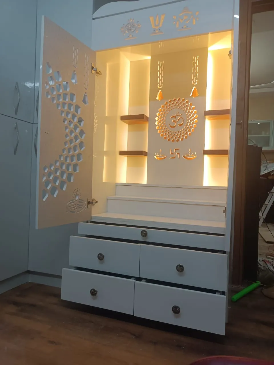 Freshly installed white mandir cabinet with CNC-cut jaali shutters, a backlit Om chakra on the rear panel flanked by warm-lit floating wood shelves