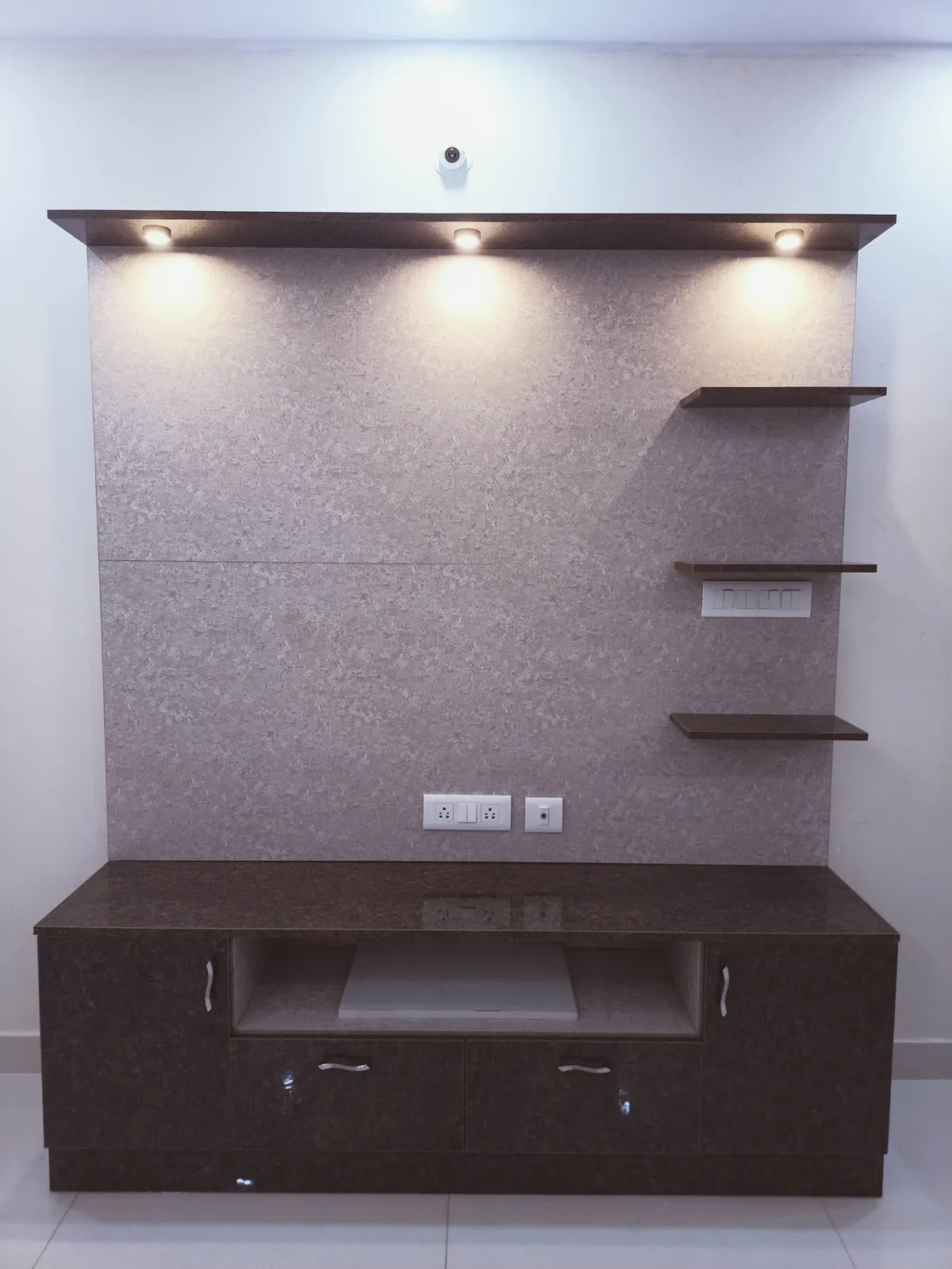 TV unit wall with a dark stone-textured backdrop, three asymmetric floating shelves, ceiling spotlighting