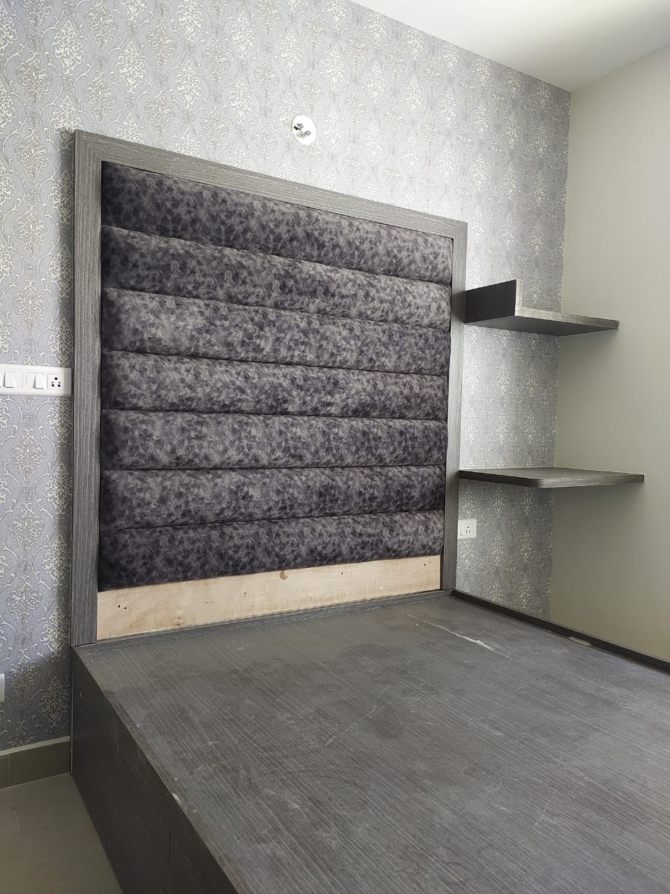 Master bedroom with a channel-tufted grey upholstered headboard on a wood-look platform bed against a grey damask wallpaper wall