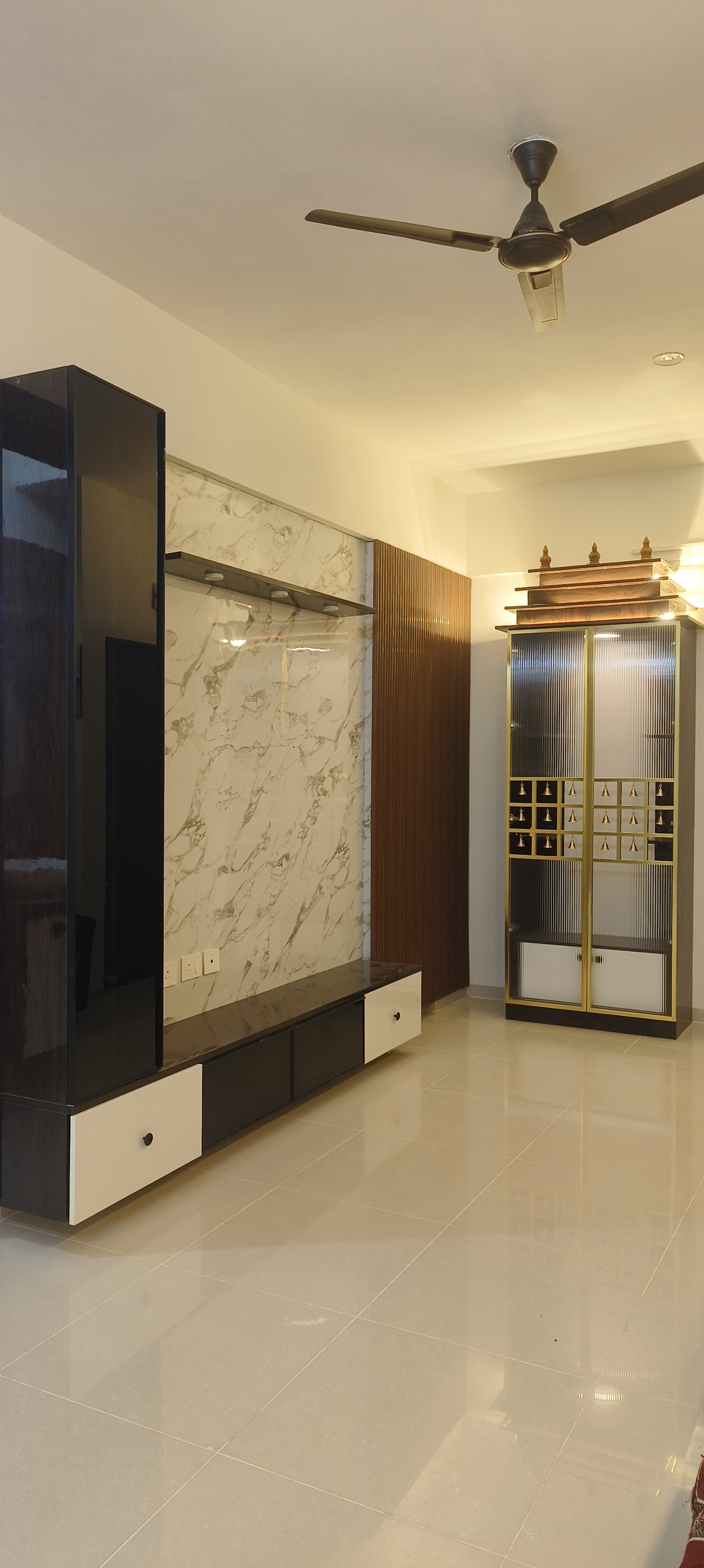 Living room corner with a black-and-marble TV feature wall beside a freestanding gold-trimmed pooja unit that has fluted glass doors and a tiered temple-style