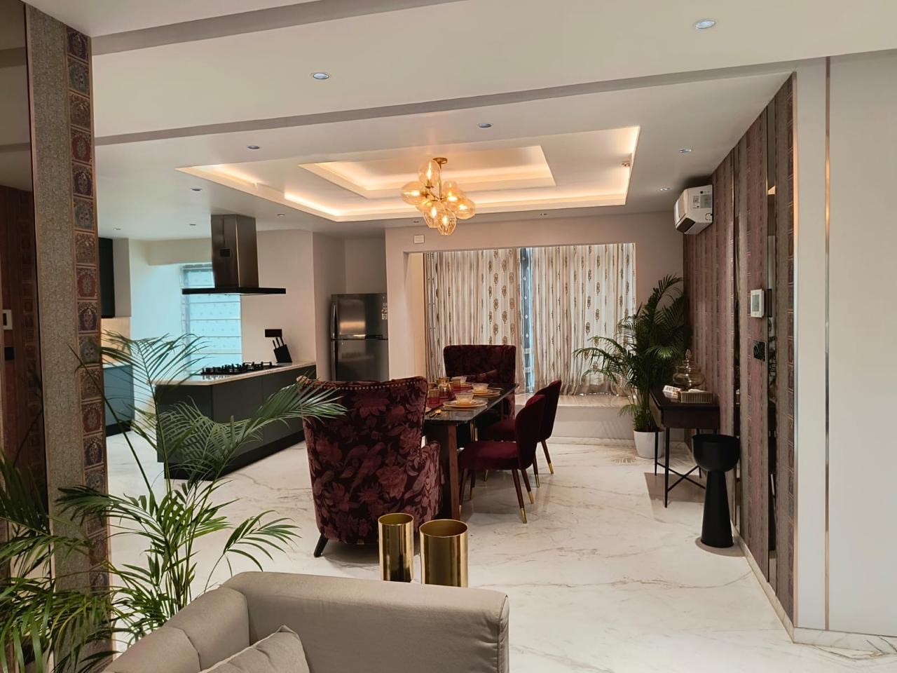 Open-plan apartment showing a dark modular kitchen at the back, a marble-top dining table with maroon floral and velvet chairs under a glass bubble chandelier