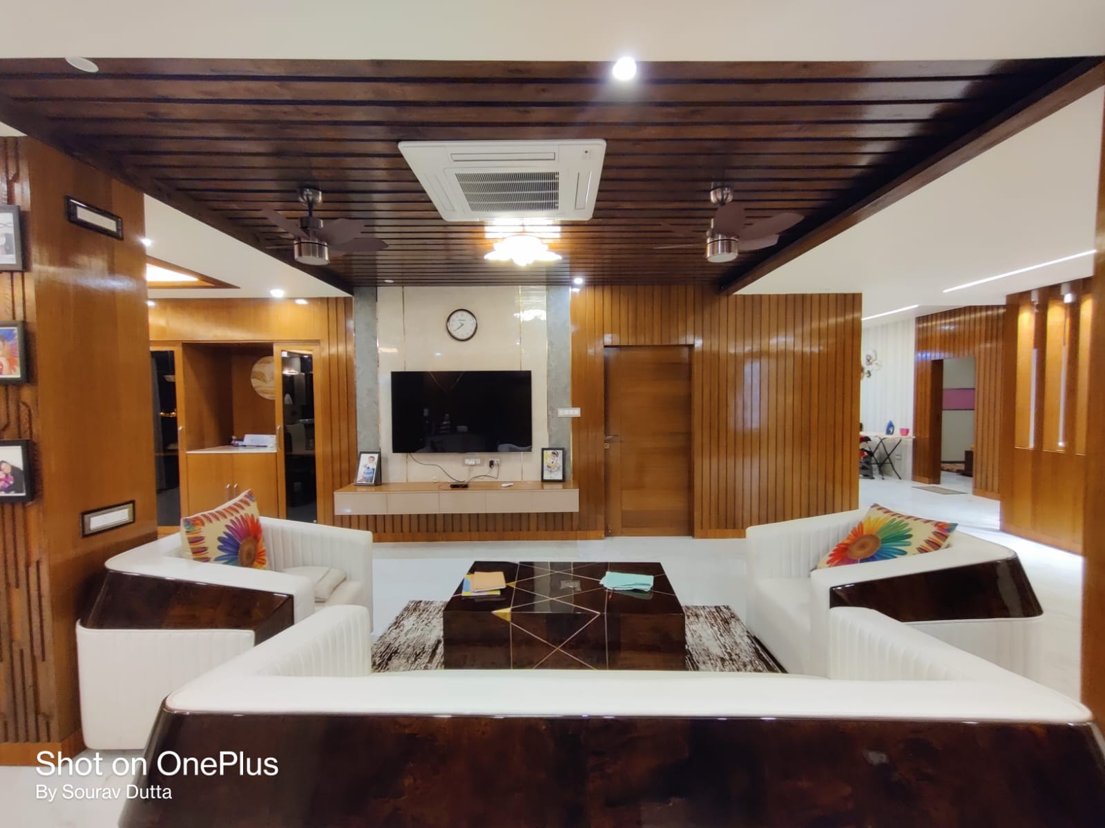 Living room with ribbed wood-panelled ceiling and walls, a floating TV console beneath a marble accent clock panel, curved white sofas