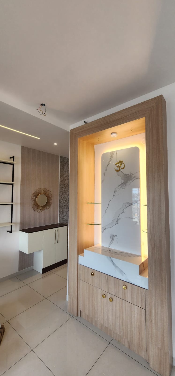 Wood-framed pooja unit with a backlit book-matched marble backdrop, a brass Om motif, glass display shelves, and storage drawers with brass knobs below.