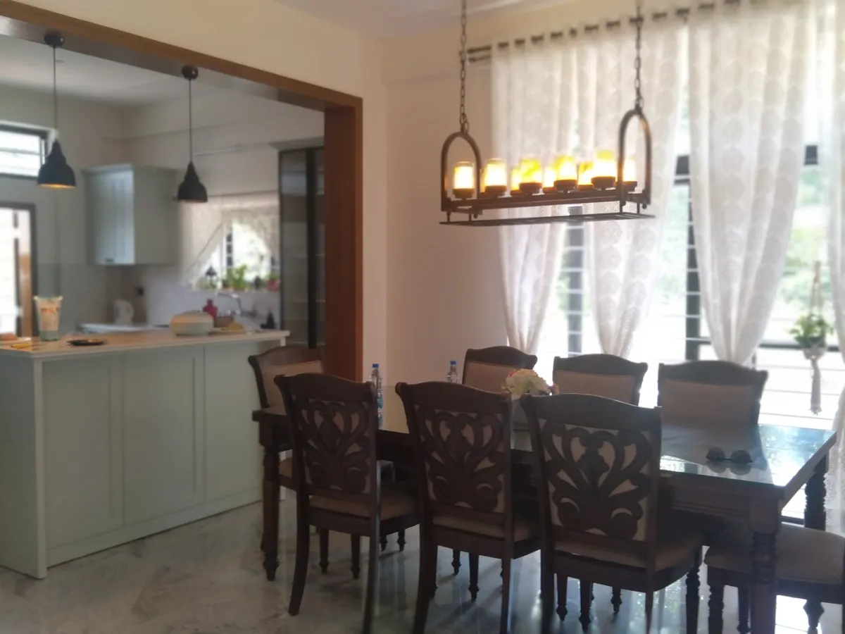 Eight-seater carved dark-wood dining set under a wrought-iron linear candle chandelier, with a sage-green breakfast-counter kitchen open behind it.