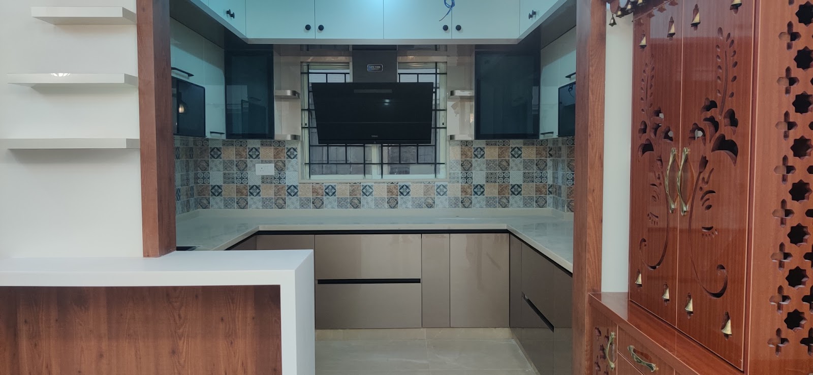 U-shaped kitchen with a patchwork mosaic-tile backsplash, a wall-mounted TV above the hob, and a carved wood pooja cabinet with brass bells visible alongside.