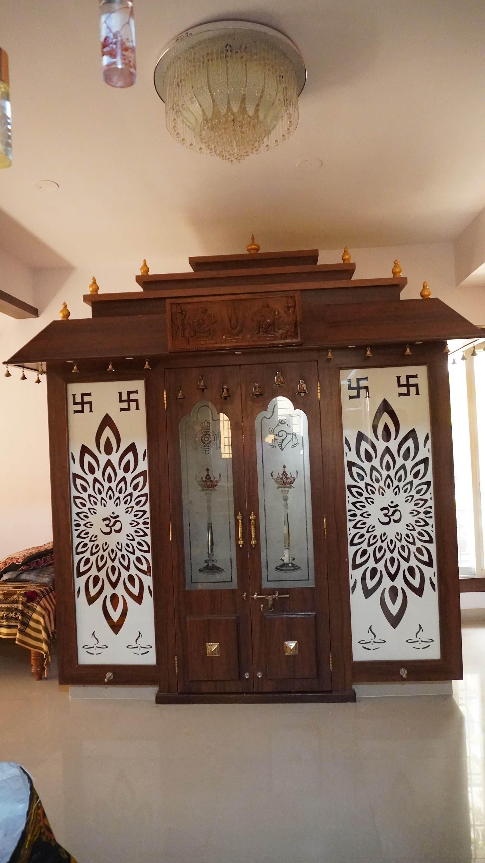 Temple-style wooden pooja mandir with a tiered dome roof, etched OM-motif glass door panels, and hanging brass bells.