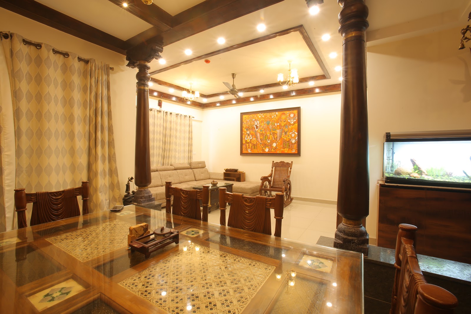 Traditional Kerala-style dining and living space with carved wooden pillars, an inlaid glass-top dining table, and a Kerala mural painting above the sofa.
